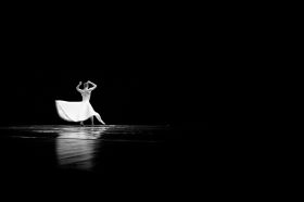 A muscular person, naked from the waist up and facing away from the camera, dances on a reflective floor. He wears a long white dress which swirls out around him and contrasts dramatically with the black background and dark, polished floor. The image illustrates On the Move, a weekly column detailing Australian arts sector appointments and resignations.