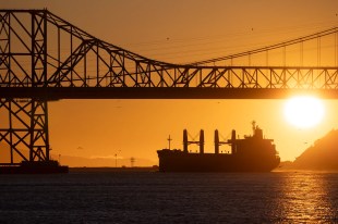 A large ship sails under a bridge at sunset. arts sector appointments.