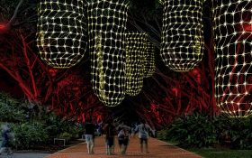 Artistic rendering of the 2024 Shine on Gimuy Artstory Light Walk, featuring yellow illuminated large-scale dilly bags hanging from tree branches. Below a group of four people are walking on the path.