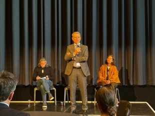 A man in a brown suit stands on stage with a microphone in front of grey/green curtains. A man and a woman are sitting either side of him. Paul Goldsmith.