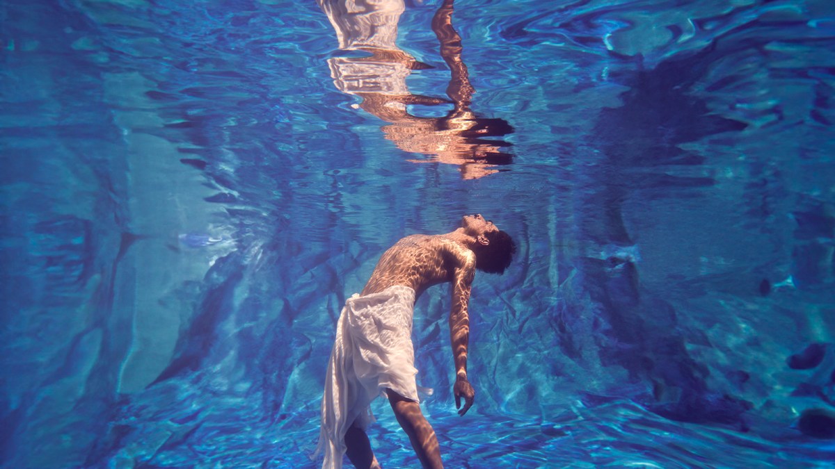 A man bare chested but wrapped in a white cloth is immersed in a body of water. His reflection can be seen above him in Horizon by Bangarra Dance Theatre.