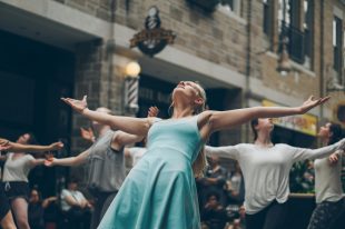 drama class. A group of young performers in an alley are all stretching their arms out and looking up in formation. The younger woman in the middle is in a greeny blue sleeveless dress.