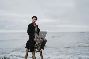 Woman sitting on a stool at waters edge, fully dressed with a laptop. Arts news.