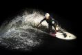 A dynamic image of a woman dressed in a back wetsuit and white helmet, riding a white surfboard. Water sprays behind her as she rides the wave. The photo illustrate's ArtsHub's weekly On the Move column about Australian arts sector appointments.