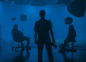 In a blue lit film studio, the silhouettes of three crew members are among the equipment. screen diversity