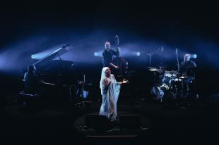 Jazzmeia Horn in white is surrounded by a band: a pianist, a double bass player and a drummer. The stage is lit in dark blue.