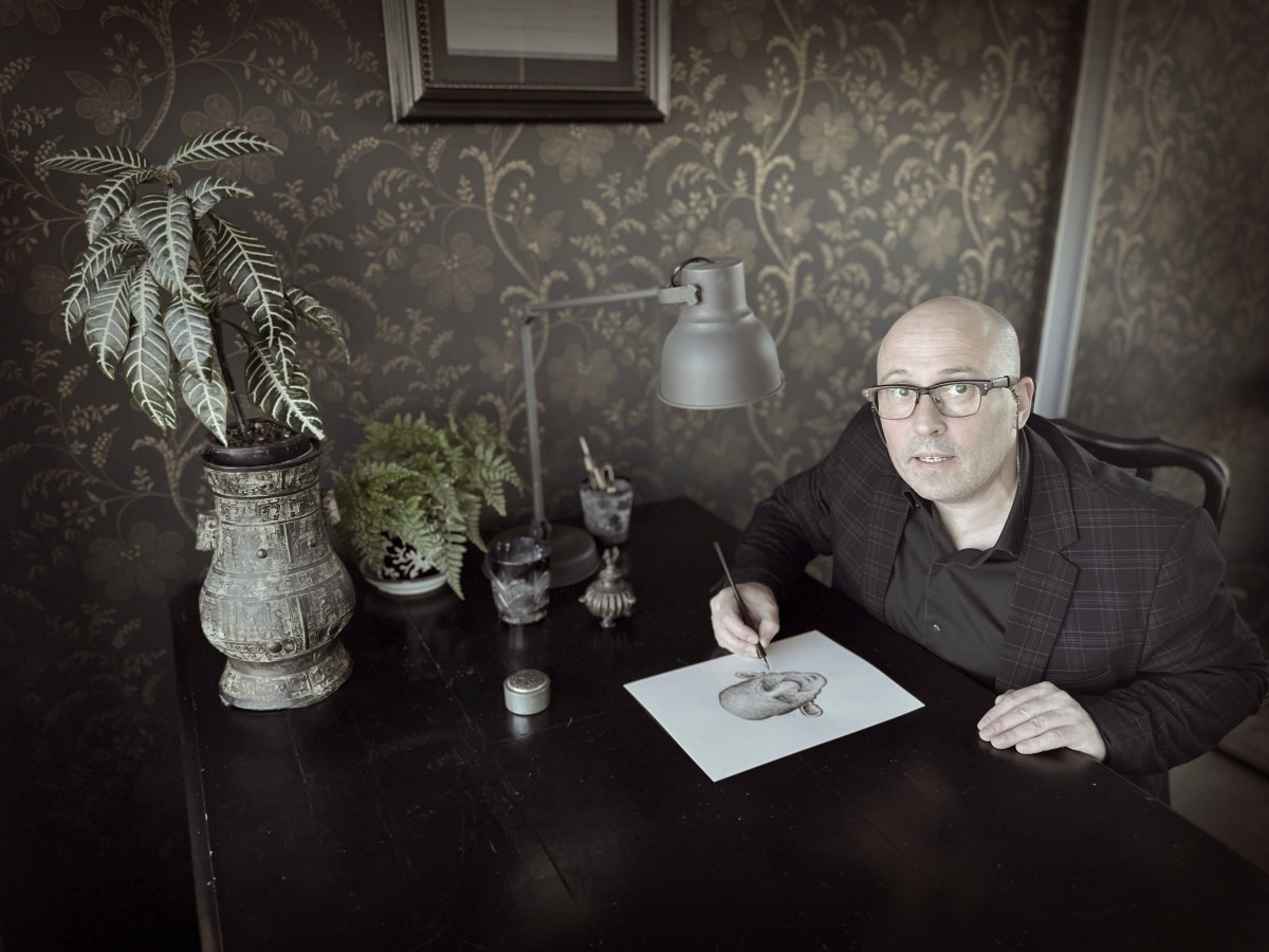 In a brown tinged room a bald headed man in glasses - Adam Elliot - sits at a table with a vase on it drawing a picture of a face. He has the pen in hand and is looking up at the camera.