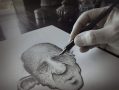 Adam Elliot at work. A grey scale close up of a face with a bald head, closed eyes and huge ears, being drawn with a pen and hand completing the drawing.