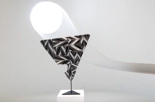 An inverted triangle with a chunk taken out of it is covered with black and white print and sitting on a small plinth. A white spotlight is shining on it.