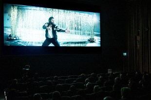 A cinema with a screen showing a man, Ben Gillies, formerly of Silverchair, standing with his legs and arms apart at a CLIPPED video session.