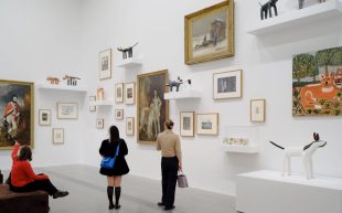 Installation view of 'Cats & Dogs' on display at The Ian Potter Centre: NGV Australia. A salon hang of staggering artworks on a white wall with three onlookers.