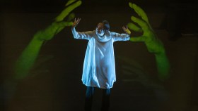 Chisato Minamimura, a woman wearing a white garment, is standing in the middle and pushing back two large disembodied hands that are lit in green in a performance of 'Scored in Silence.'