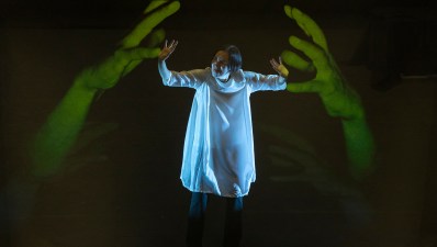 Chisato Minamimura, a woman wearing a white garment, is standing in the middle and pushing back two large disembodied hands that are lit in green in a performance of 'Scored in Silence.'