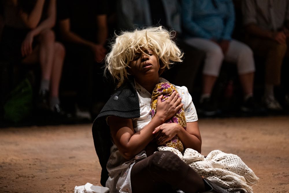 A scene from the upcoming 2025 Sydney Festival production 'Dark Noon'. A black-skinned South African person wearing a bedraggled blonde wig sits on a dirt stage and cradles what appears to be a baby wrapped in blankets to their chest.