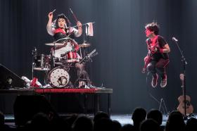 A woman is drumming on a raised stage. Besides her on the right is a man with a guitar mid leap in the air. They are Astrid & Otto, and dressed in shades of black and red.