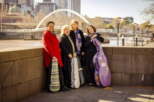 Four women in winter coats lean against a wall on the banks of the river Yarra. With Melbourne's Flinders Street station on the other side of the river. They all have instrument cases - two violins, one viola and a cello. Flinders Quartet