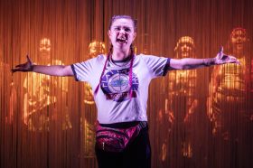 A young girl with her arms outstretched and wearing a tshirt that says 'Flat Earth Society' , is standing in front of a scrim, behind which stands shadowy figures.