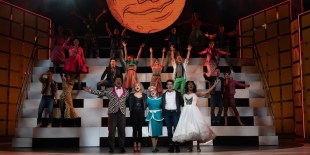 The cast of 'Grease'. They are standing with their hands in the air. A white platform of stairs behind them.