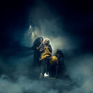 production of 'Macbeth', which opens the company's 2025 season. Three older women, playing witches, pose in front of a smoky and dramatically lit background.