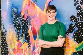 Amy MacMahon, the Queensland Greens MP for South Brisbane and spokesperson for the arts, stands in front of a mural painted by Thistle Pearl @thistlesanctuary. Amy is a fair-skinned woman with short brown hair and glasses and wearing a green t-shirt. She stands in front of a vibrantly coloured piece of street art.