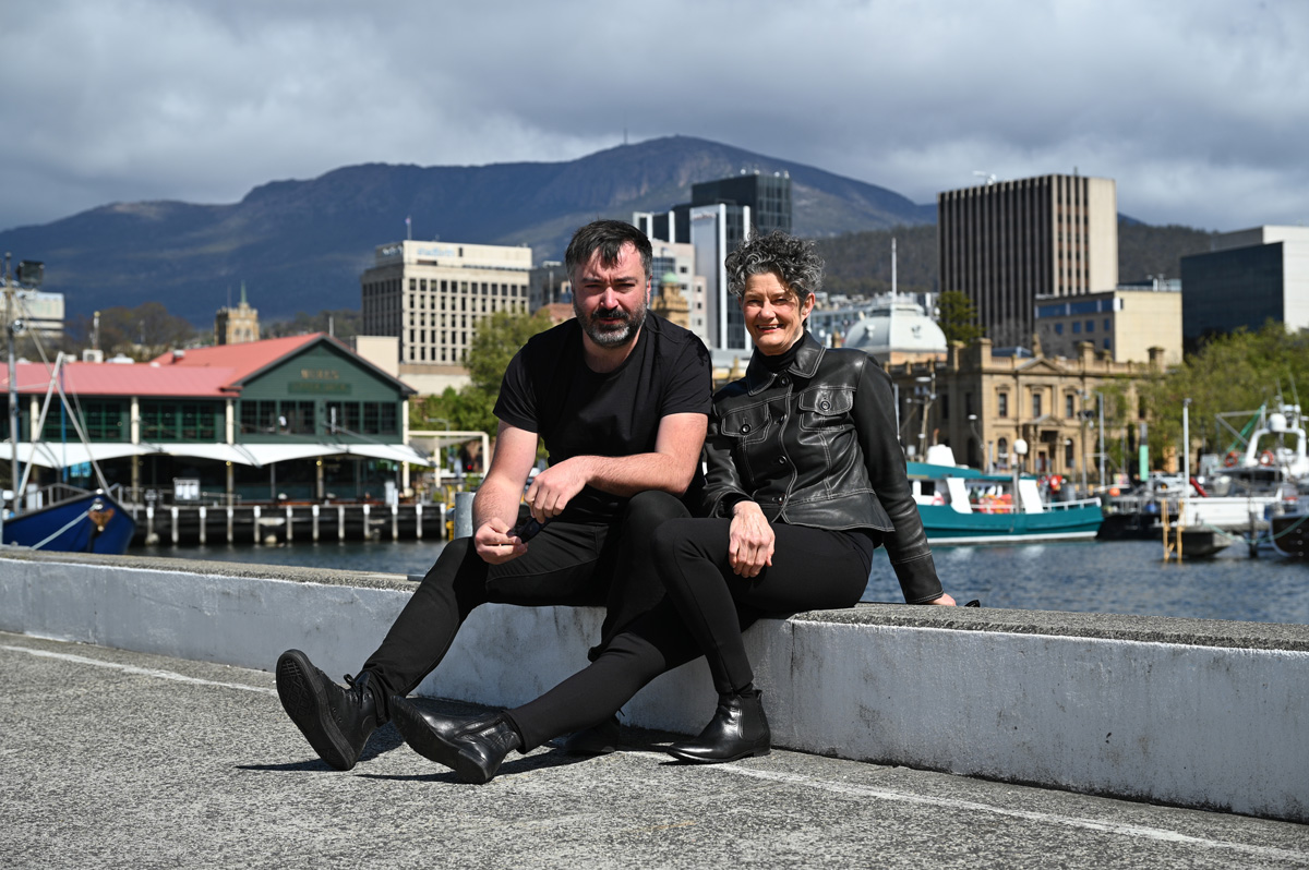 Travis Tiddy and Denise Robinson are the new co-Creative Directors of the biennial exhibition Hobart Current. On the Move: Australian arts sector appointments. A bearded man and a dark-haired woman, both dressed in black, sit at Hobart's waterfront with the city and kunanyi Mt Wellington visible behind them.