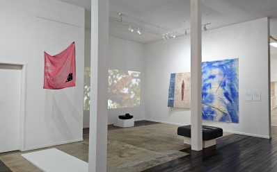 ‘skeletons’ by Judy Watson, installation view at Umbrella Studio Contemporary Arts, Townsville. A white-walled gallery with two large columns in the middle. Several works are on display including a sheer red banner on the left and a video projection showing landscape in the centre.