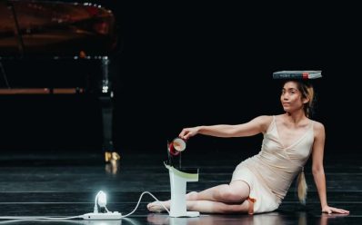 A performer on stage who is of Asian descent with a female-appearing body, wearing a nude slip. The performer is sitting on the side, holding a cup on top of the coffee machine and balancing a book on their head.