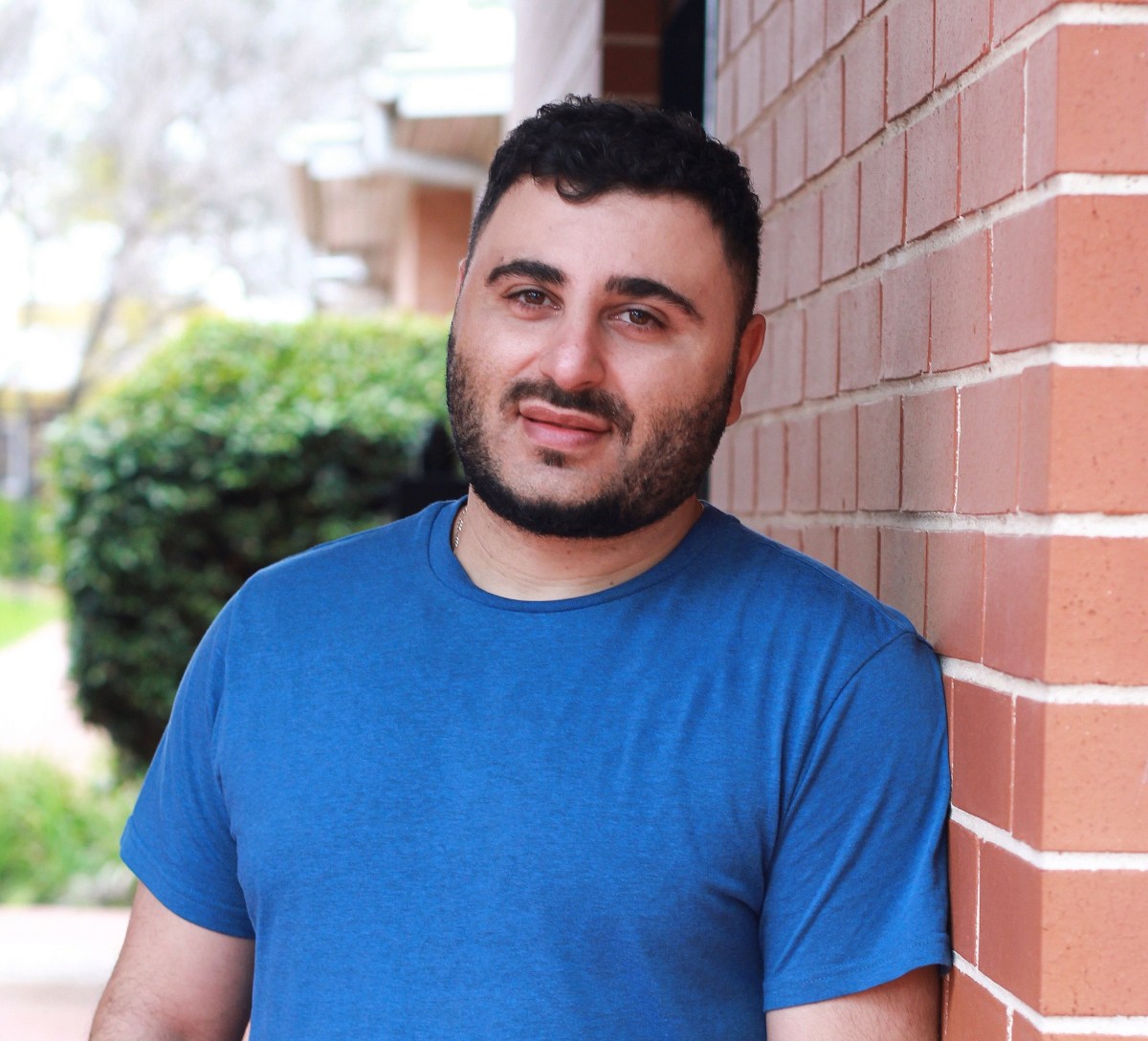 James Elazzi. A Young Man In A Blue T Shirt With A Dark Beard And Moustache Leans Against A Wall, With A Hedge In The Background