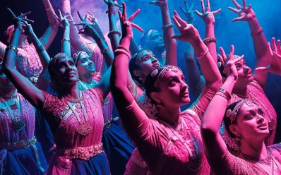 A group of Indian dancers in pink and purple are dancing with their hands above their heads.