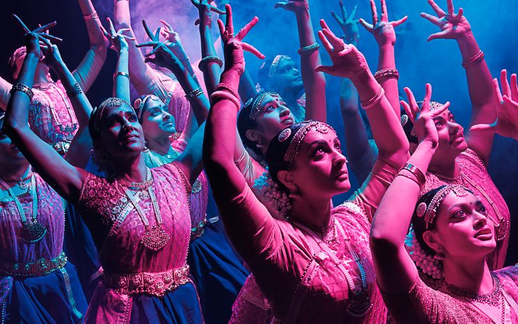 A group of Indian dancers in pink and purple are dancing with their hands above their heads.