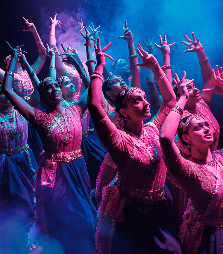 A group of Indian dancers in pink and purple are dancing with their hands above their heads.