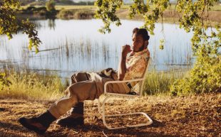 Leon Bridges will performed as part of the 24th A Day On The Green in 2025. A black man sitting on an outdoor chair next to a lake.