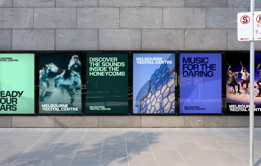 A grey wall and pavement, on the wall are a series of posters side by side advertising concerts at the Melbourne Recital Centre (MRC) with lines like Discover the Sounds inside the Honeycomb and Music for the Daring.