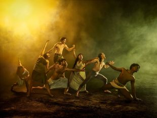 A publicity image for 'Troy', one of seven productions in Malthouse Theatre's 2025 season. A multi-racial cast of seven, including men in skirts and women in tunics reminiscent of ancient Greece, link hands as they struggle across a dramatically lit and smoky battlefield.