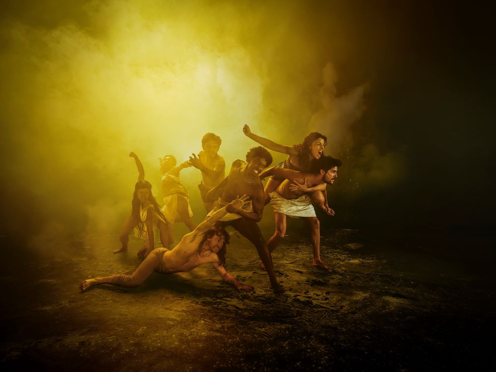 A publicity image for ‘Troy’, one of the productions in Malthouse Theatre’s 2025 season. On a dirt floor, seven characters dressed in togas and loincloths emerge from a yellow fog, their bodies struggling, sprawled and contorted.