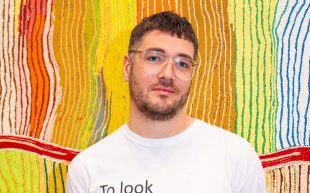 Myles Russell-Cook to lead ACCA. A young First Nations man with pale skin, short cropped brown hair and beard, wearing large clear frame glasses and a t-shirt with the text 'To look/without fear', standing in front of a colourful painting with dot work.