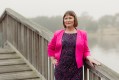 The ACT's Shadow Minister for Arts Nicole Lawder, discusses arts policy in the lead-up to the ACT election. Nicole wears a bright pink jacket and has neat brown hair; she stands on a wooden bridge over a misty lake with her right hand on her hip and her left hand on the bridge railing.