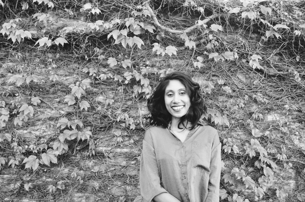 Nicole Reddy. Photo Is A Black And White Image Of A Woman In Front Of A Wall With Ivy Leaves On It. She Is Wearing An Open Necked Shirt And Has Shoulder Length Dark Hair