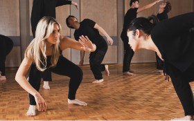 Auditions for the NIDA Open 2025 Studios program open in December. The photo shows a group of young people in theatre blacks in a rehearsal studio, with the focus on two young women crouched on the floor and gesturing to one another expressively.