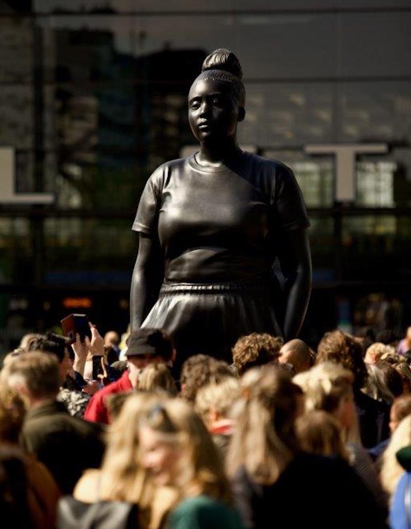 oversized sculpture of a young woman in everyday dress, with crowd taking photos of it. Thomas J Price