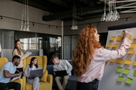 a group of young people in a room comparing notes on computers. a red-haired woman is in the foreground putting sticky notes on a whiteboard. writers' room