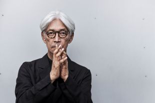 The late Ryuichi Sakamoto will perform in a virtual performance at Asia TOPA 2025. A white-haired Japanese man in dark glasses and a black shirt poses for the camera with his raised hands held to his lips.