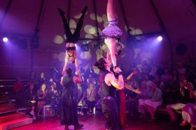 A small circus tent with an audience watching four acrobats, with two men at the bottom balancing upside down women on their hands. So Soiree