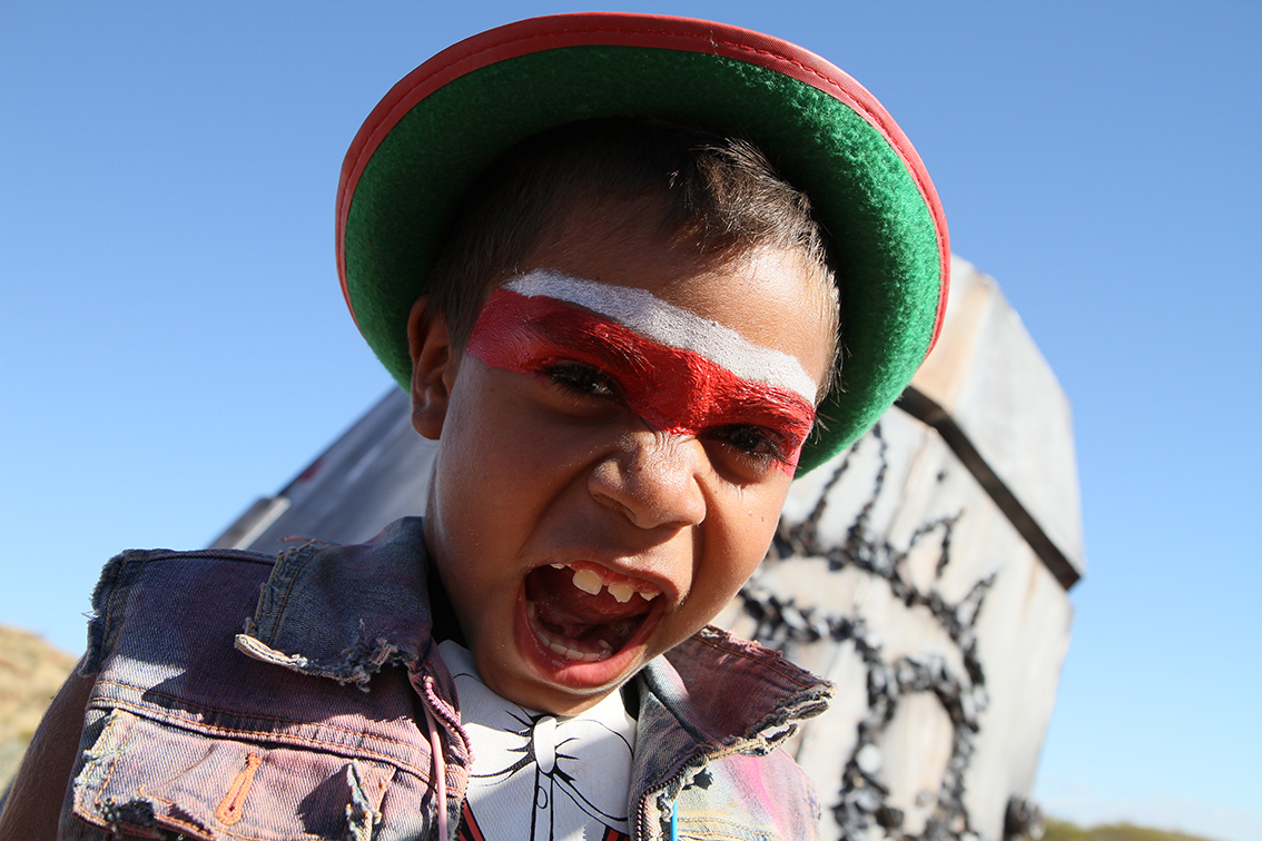 A young First Nations boy with red and white striped face paint across his brow and a green brimmed hat, and a ragged denim sleeveless jacket roars at the camera. The Future and Other Fictions. the Love Punks, Sutu