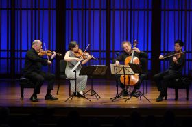 Colarado's Takács Quartet will collaborate with Australian actor Angie Milliken in Musica Viva's 2025 season. One woman and three men, the members of a string quartet, play on a wooden stage against a blue-lit backdrop.