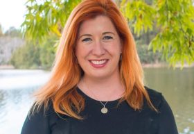 Tara Cheyne, ACT Minister for Arts, Culture and Creative Economy. A fair-skinned woman with long red hair smiles at the camera. She wears a black shirt and a silver necklace and is standing with a lake and trees visible behind her.