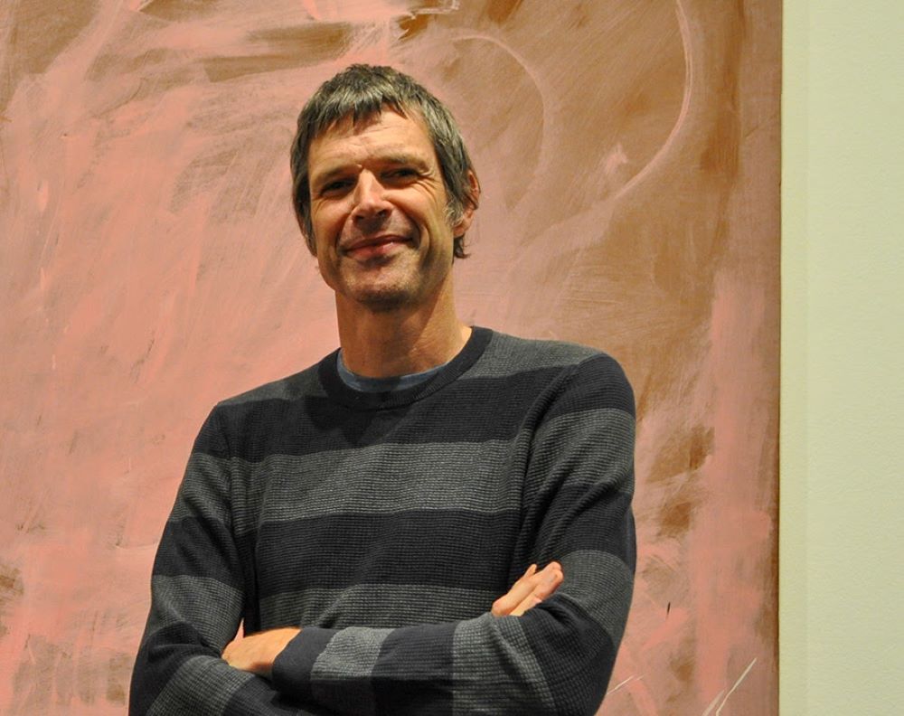 Anthony (Tony) Oates, the new Director of Frill Hall Gallery, poses with his arms folded. He wears a striped gray jumper and is standing in front of a painting.