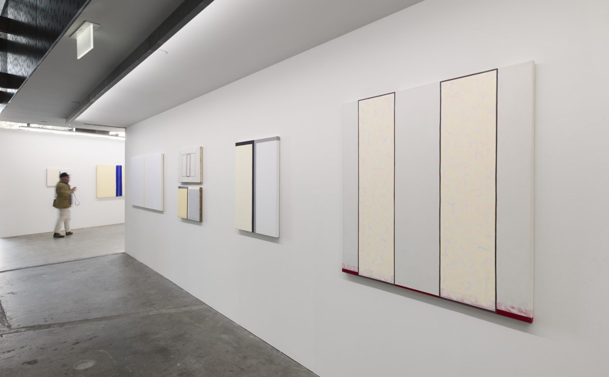 A white walled gallery space showing a series of large minimalist abstract paintings that have white, grey and cream colours.