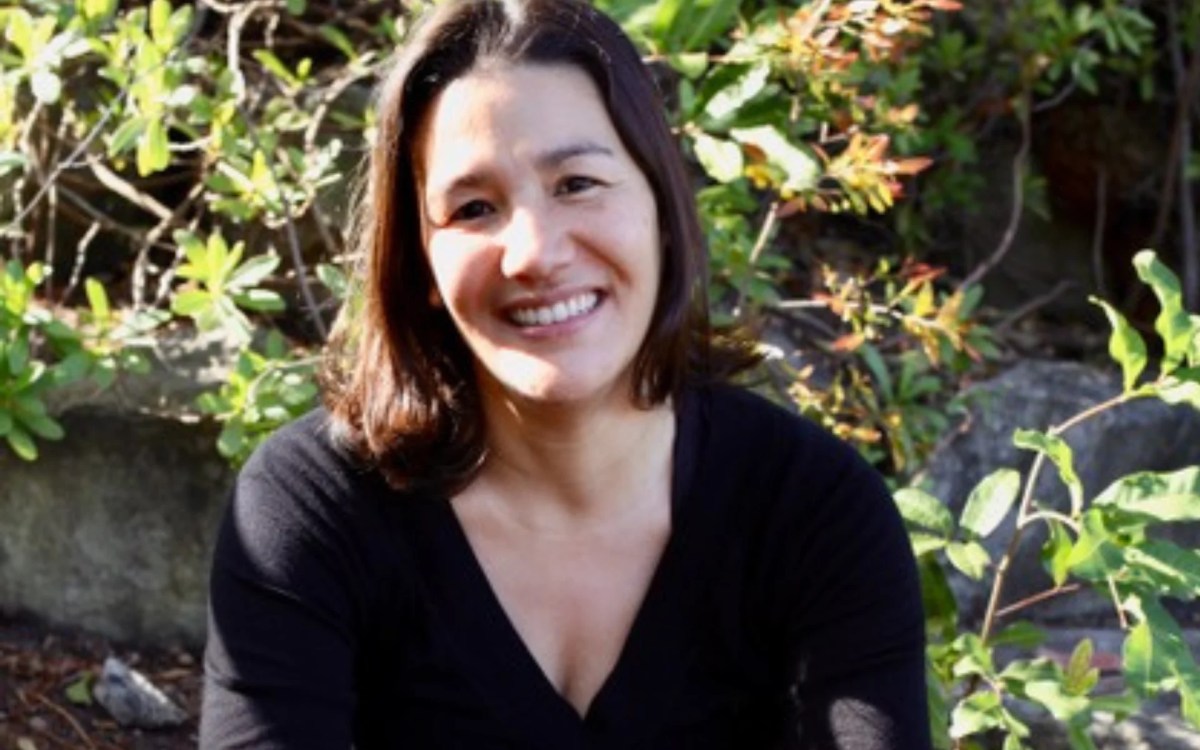 Clare Atkins. Photo Is.a Young Woman In Front Of A Shrub Wearing A Black V Neck Jumper With A Dark Shoulder Length Bob.