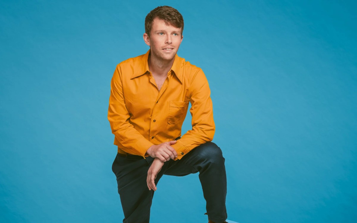 Jack Druce. Young Red Haired Man In An Orange Shirt And Dark Trousers Against A Blue Backdrop.